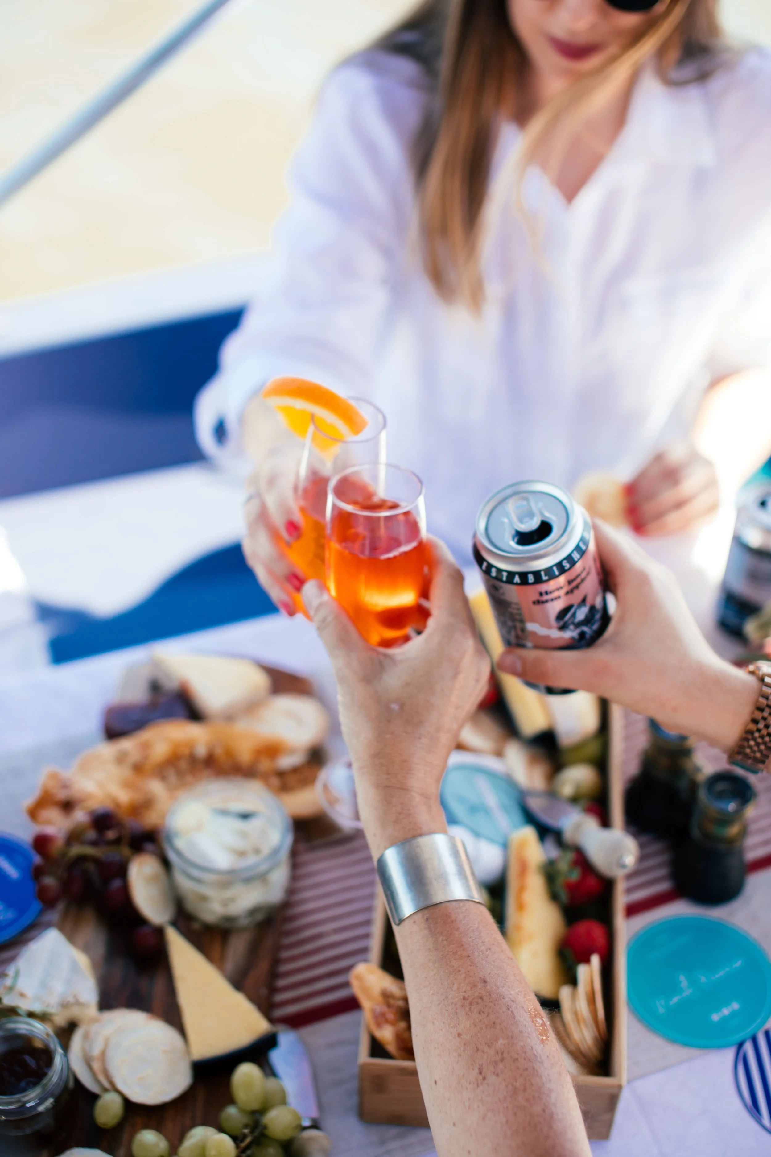 People toasting with glasses of orange-colored drinks at a table filled with food, including cheese, bread, grapes, and dips.