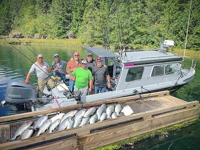 Seven people standing on a boat with a wooden edge, posing with a catch of fish laid out on the edge. The boat is on a body of water with tree-lined shore in the background.