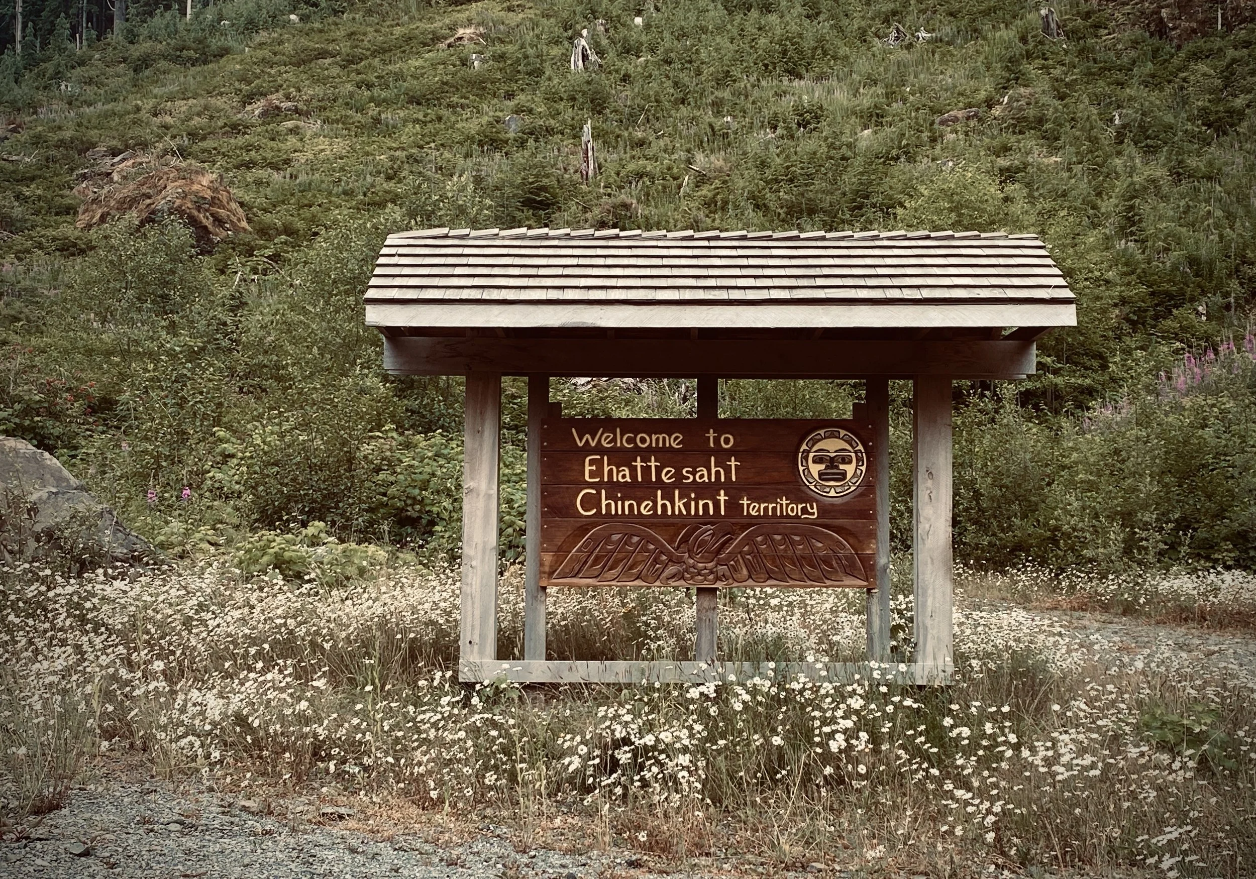 A wooden welcome sign in front of a lush green forested hillside with wildflowers at the base, indicating Chattesht, Chinhkint territory.