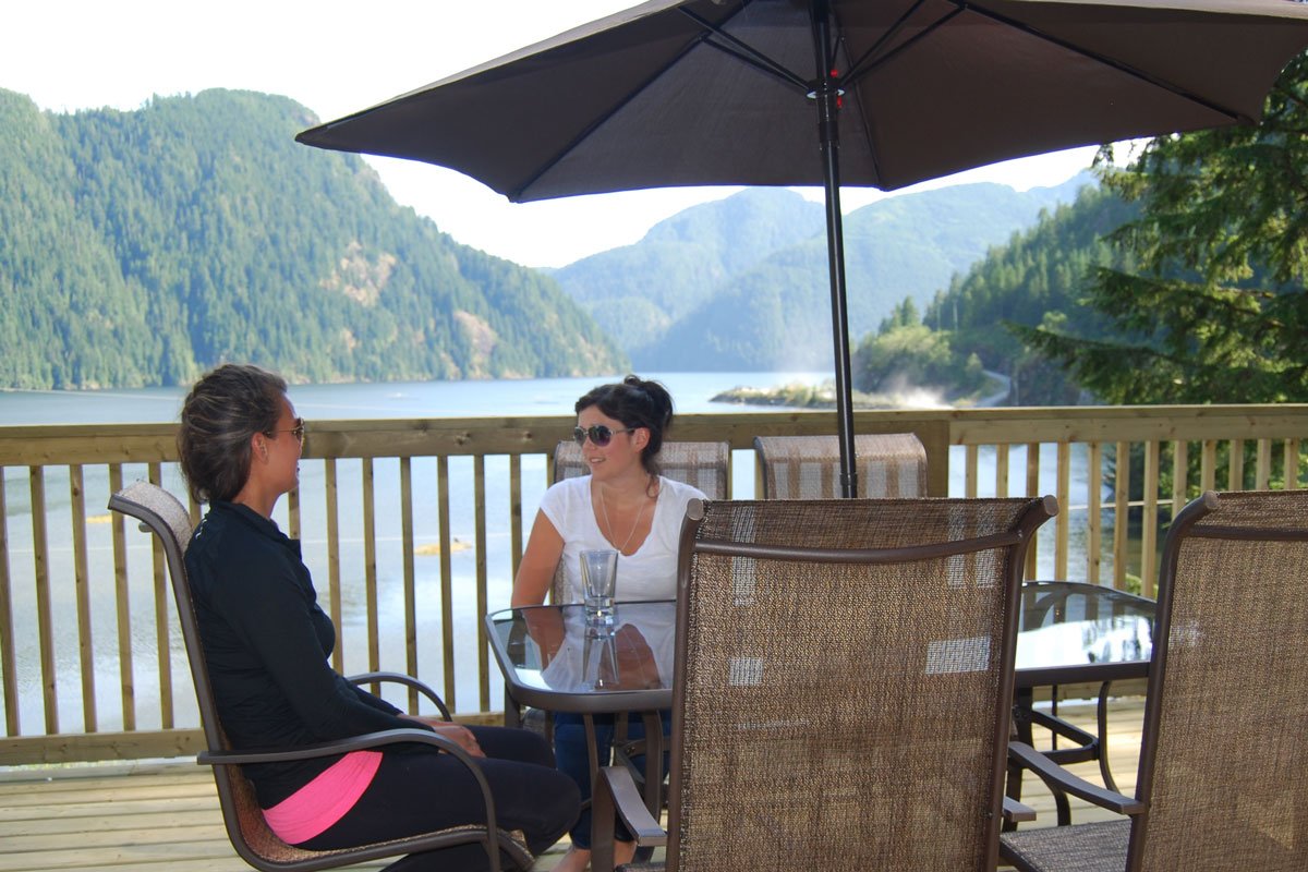 Two women sitting at a glass table on a wooden deck with a lake and mountains in the background, shaded by a large umbrella.