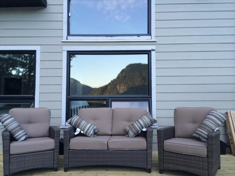 Outdoor patio furniture set with two armchairs and a three-seater sofa with striped pillows, situated on a wooden deck in front of a house with three large windows reflecting a mountainous landscape.