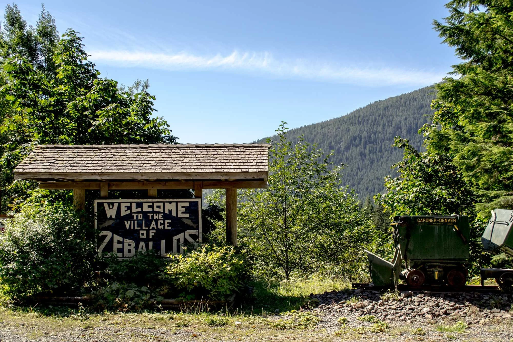 A wooden signboard reading 'Welcome to the Village of Zephyr' is situated among green trees and shrubs, with a mountain covered in pine trees in the background. There is a small building with a shingled roof and a piece of construction equipment or a small vehicle nearby.