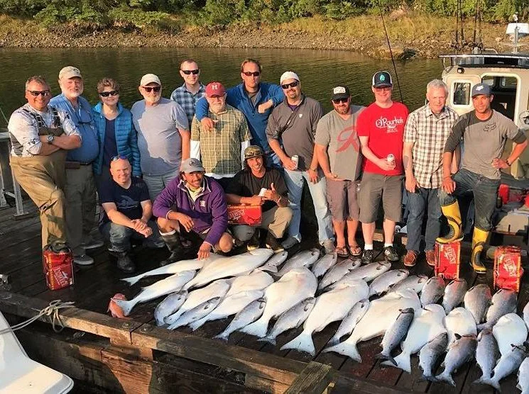 Group of people on a wooden dock by a body of water, displaying a large catch of fish laid out in front of them, with trees in the background.