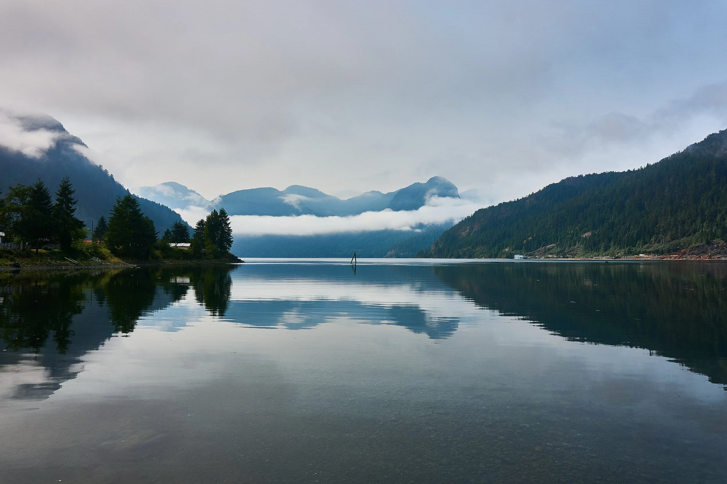 Calm lake reflecting surrounding green forested mountains under a cloudy sky.