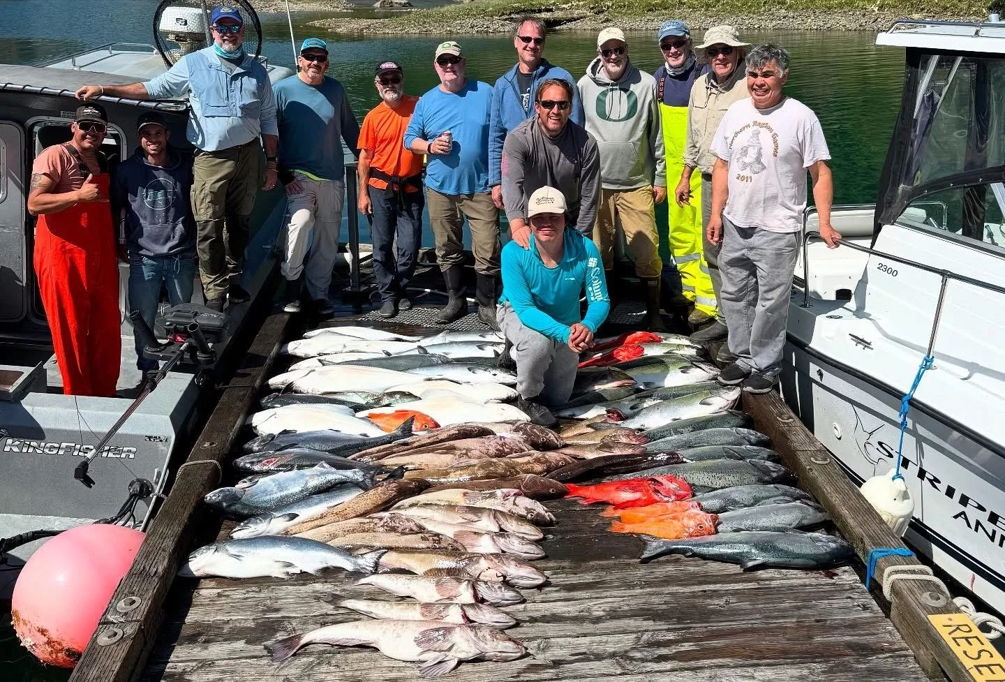 Group of people on a dock with fishing boats, displaying a large catch of various fish species laid out on the wood planks, with water and shoreline in the background.