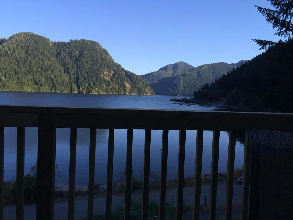 A view of a lake surrounded by green mountains, seen from a balcony with a wooden railing and a chair.