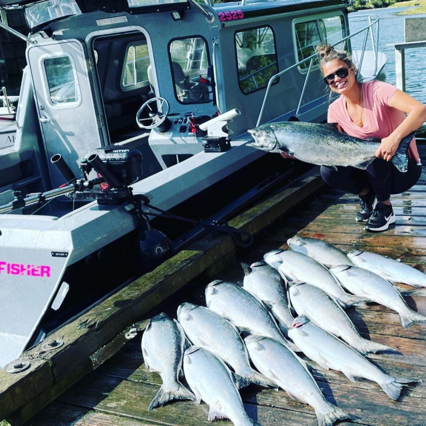 A woman on a dock holding a large fish, with several other fish laid out on the wooden dock, and a boat in the background.