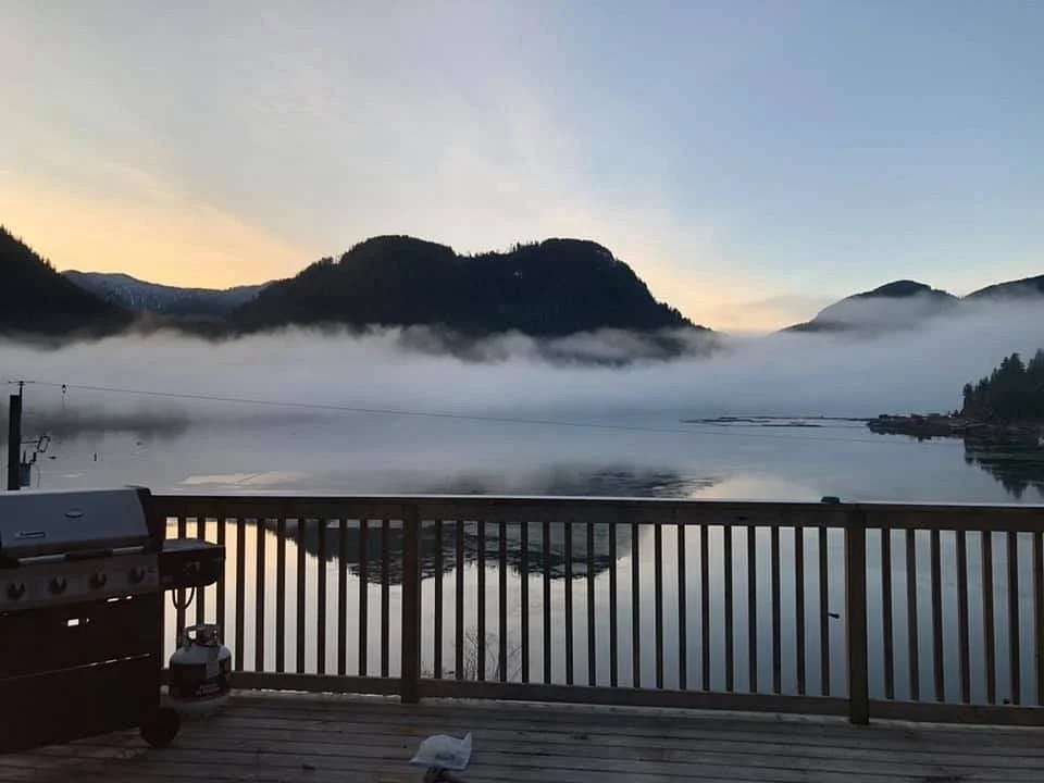 A serene lake scene at dawn with mist over the water, surrounded by mountains, viewed from a wooden deck with a grill and propane tank.
