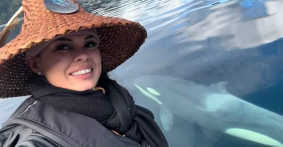 woman wearing a wide-brimmed straw hat and black jacket, smiling near a body of water with a large shark swimming close underneath.