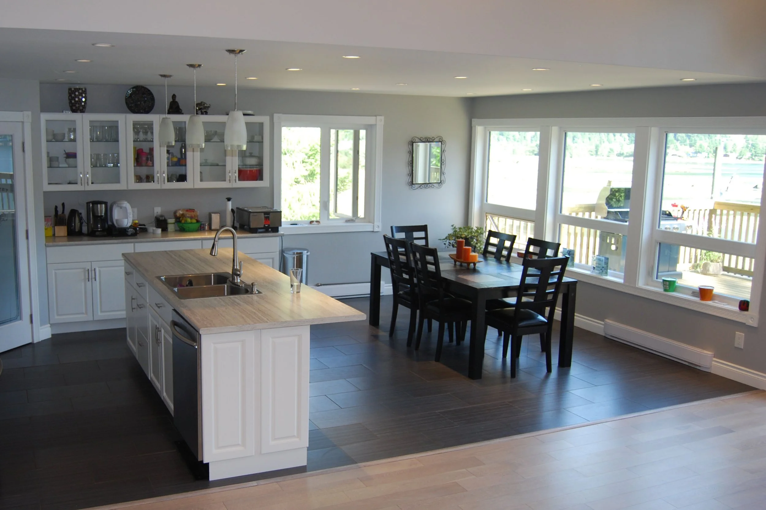 A modern kitchen and dining area with large windows, white cabinetry, a kitchen island, black dining chairs, and a view of outdoor scenery.