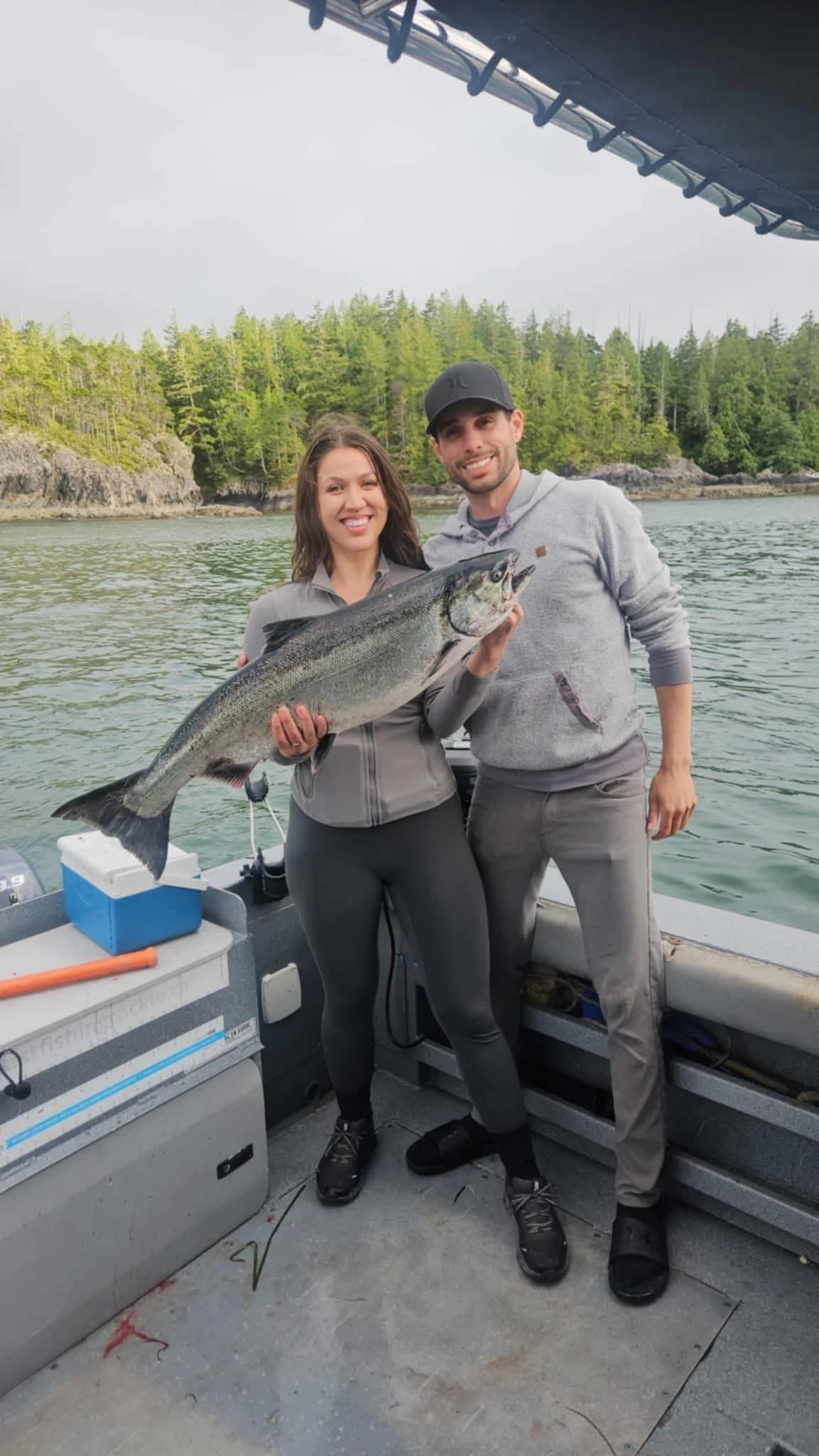 Two people on a boat holding a large fish, with a forested shoreline in the background and a cloudy sky overhead.