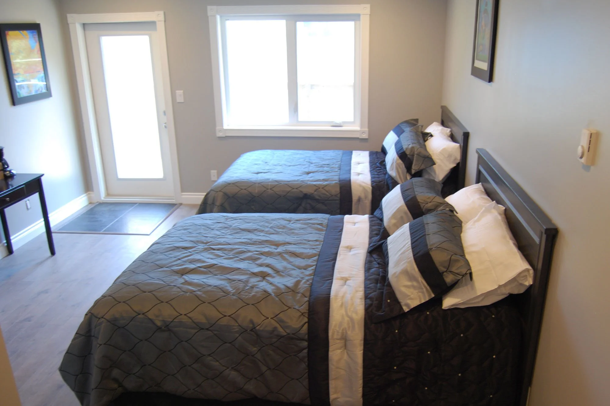 A small bedroom with two twin beds with black, gray, and white bedding, dark wood headboards, and pillows. There is a large window with plantation shutters, a door with frosted glass, a small black nightstand, framed colorful artwork on the walls, an