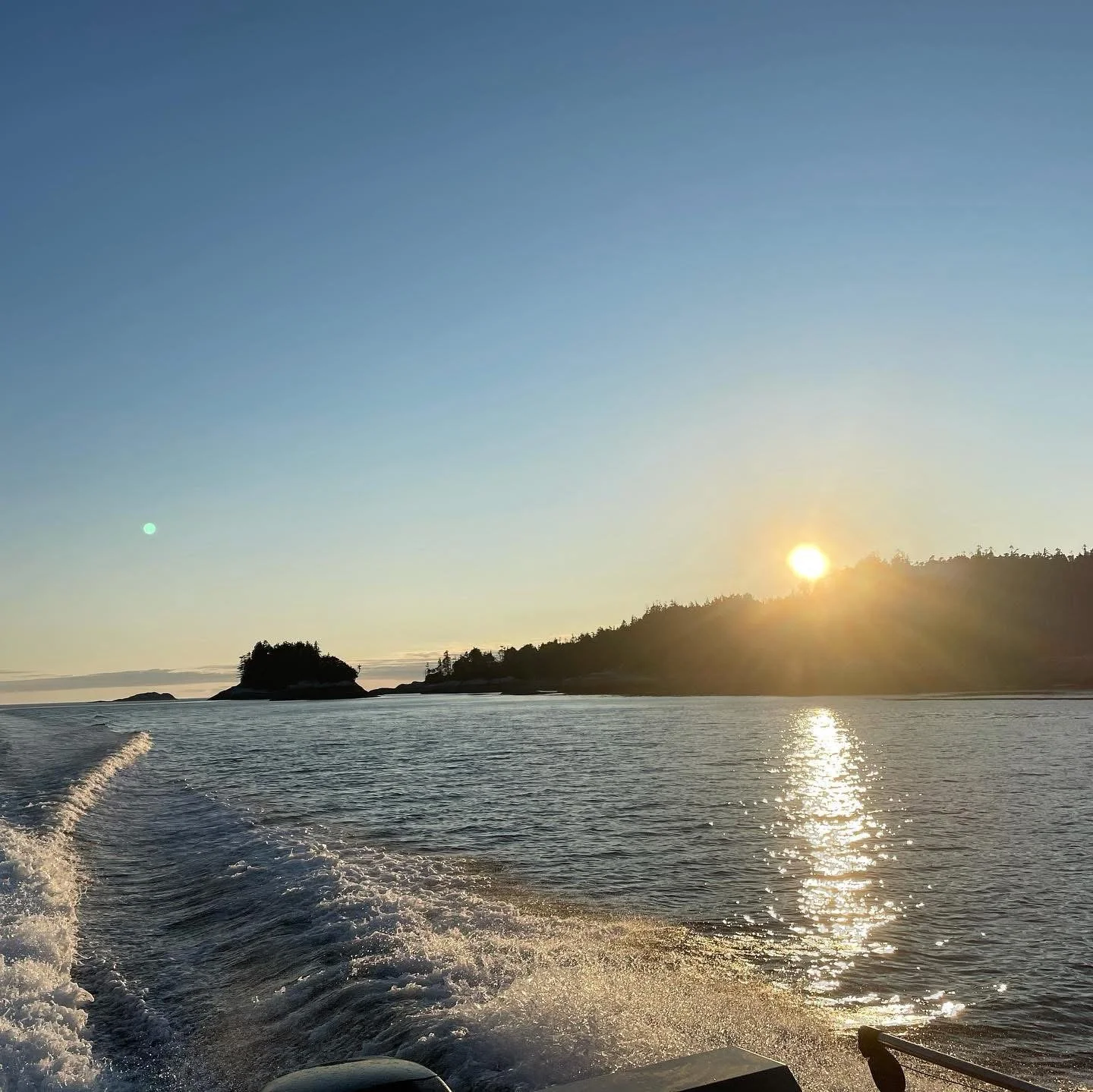 Sunset over a calm ocean with small islands and a forested shoreline, seen from a boat.