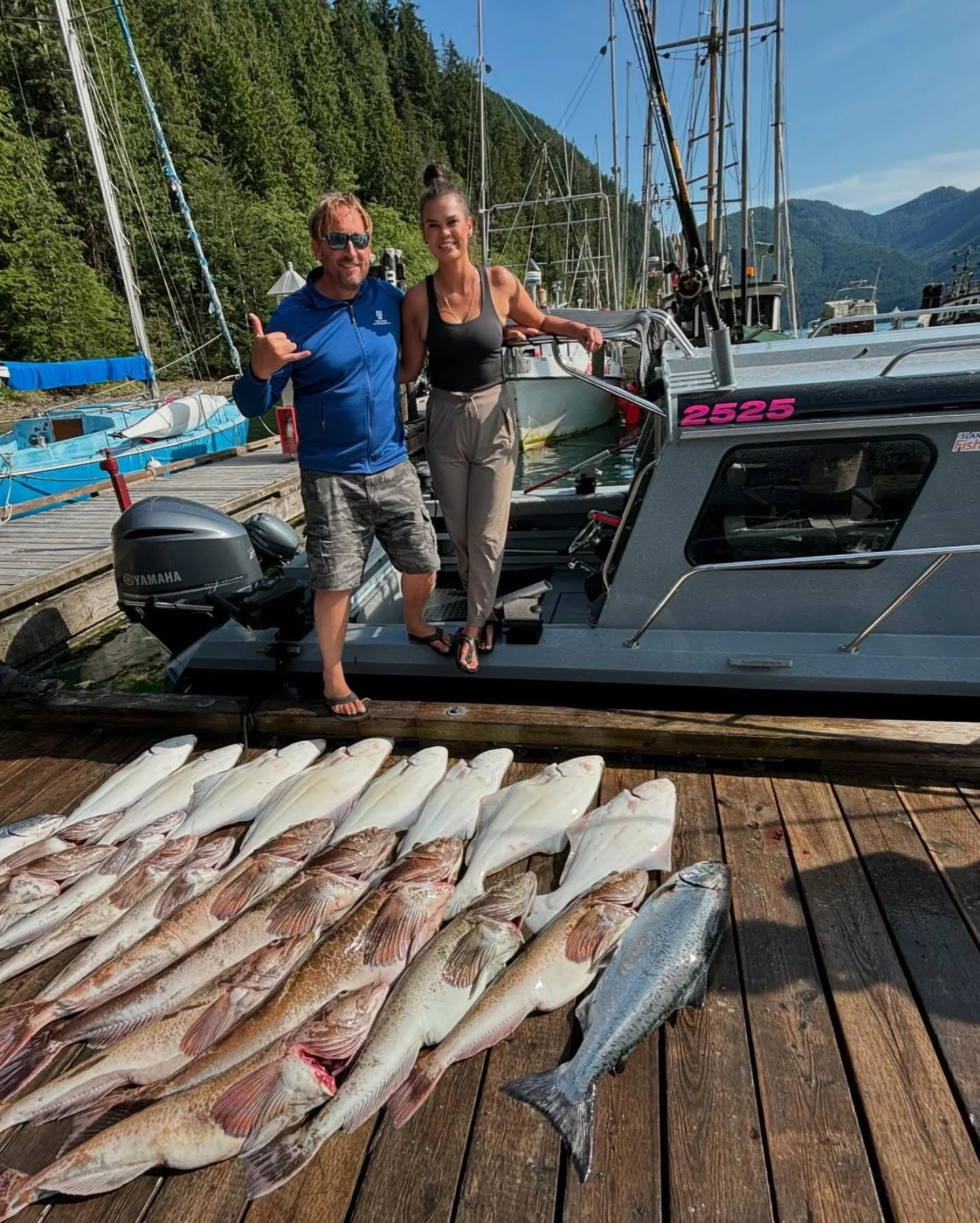 Two people standing on a dock next to a boat with a display of fish laid out in front of them, mountains and other boats in the background.