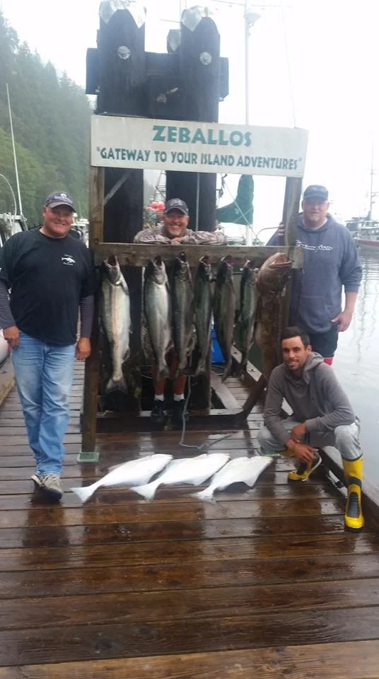 Four men on a wooden dock display caught fish, with some hanging from a wooden frame and others lying on the dock, near a body of water with boats in the background. A sign reads 'Zeballos, Gateway to Your Island Adventures'.