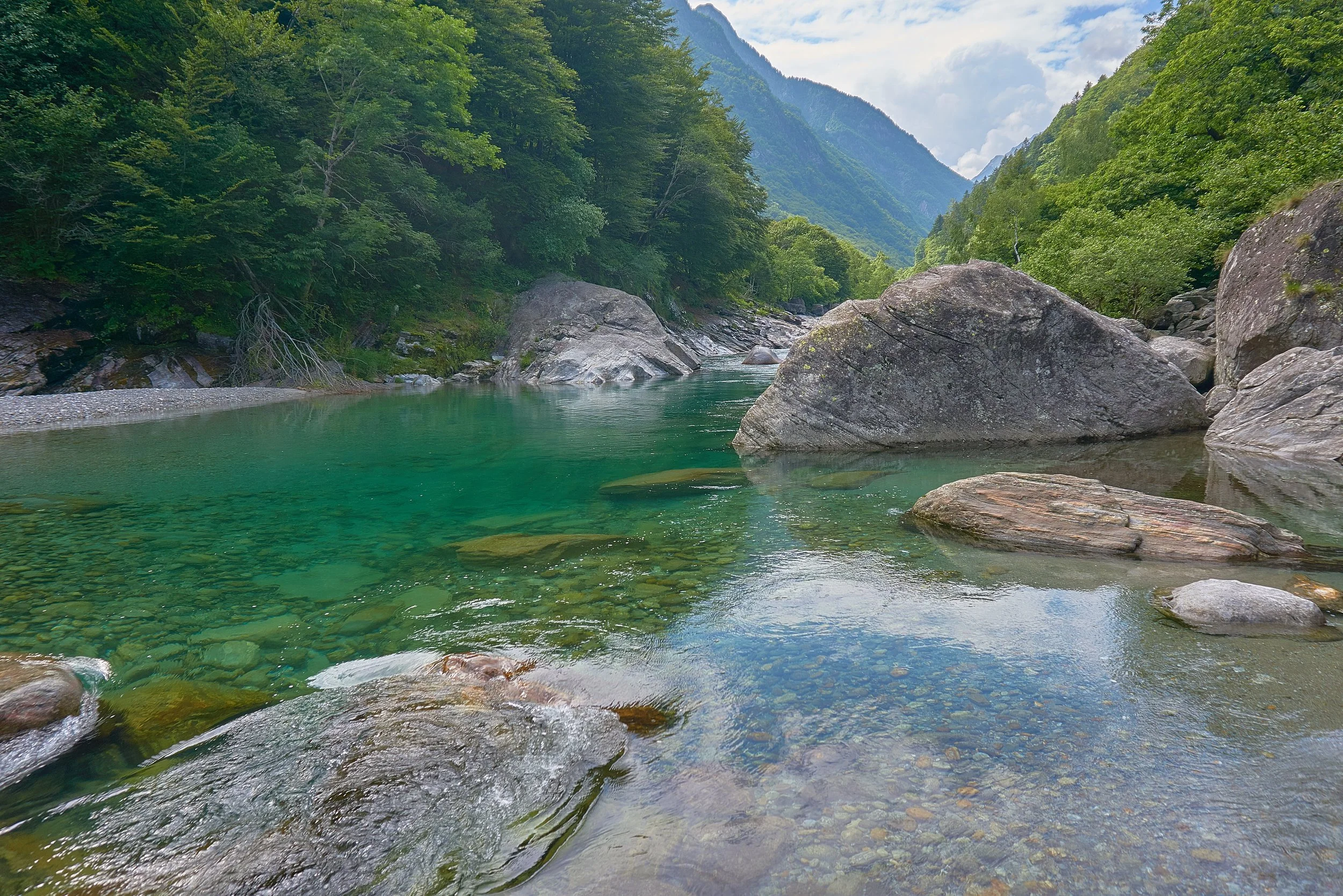 A clear green river flowing through a lush, green forested landscape with large rocks and mountains in the background.