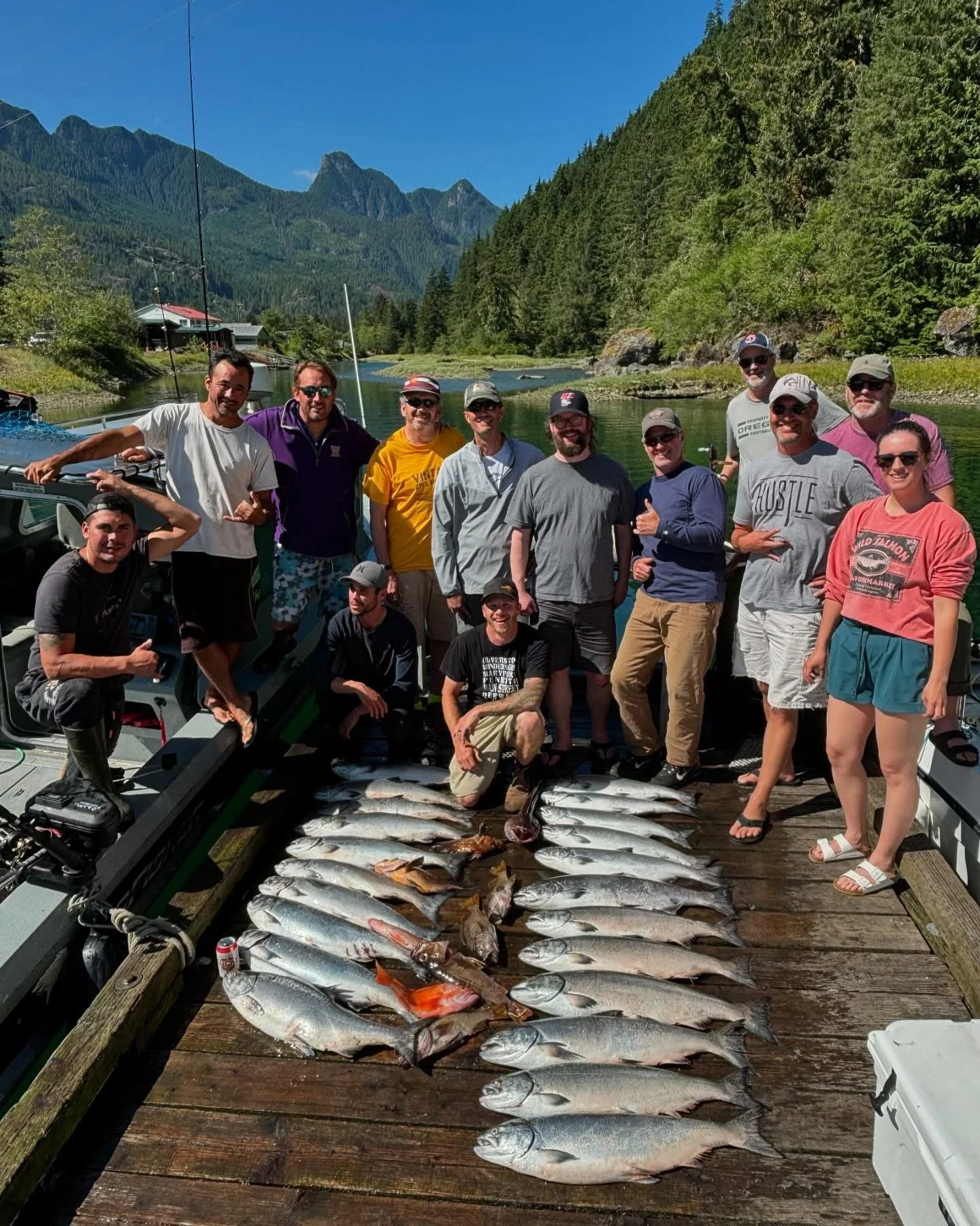Group of people on a dock in a mountainous area with trees and water, posing with a large catch of fish laid out on the dock in front of them.