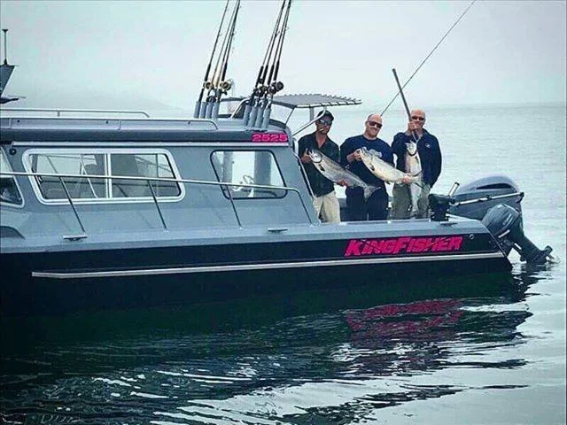Three men on a boat holding fishing rods and a large fish, with the boat labeled 'KingFisher' and the ocean in the background.