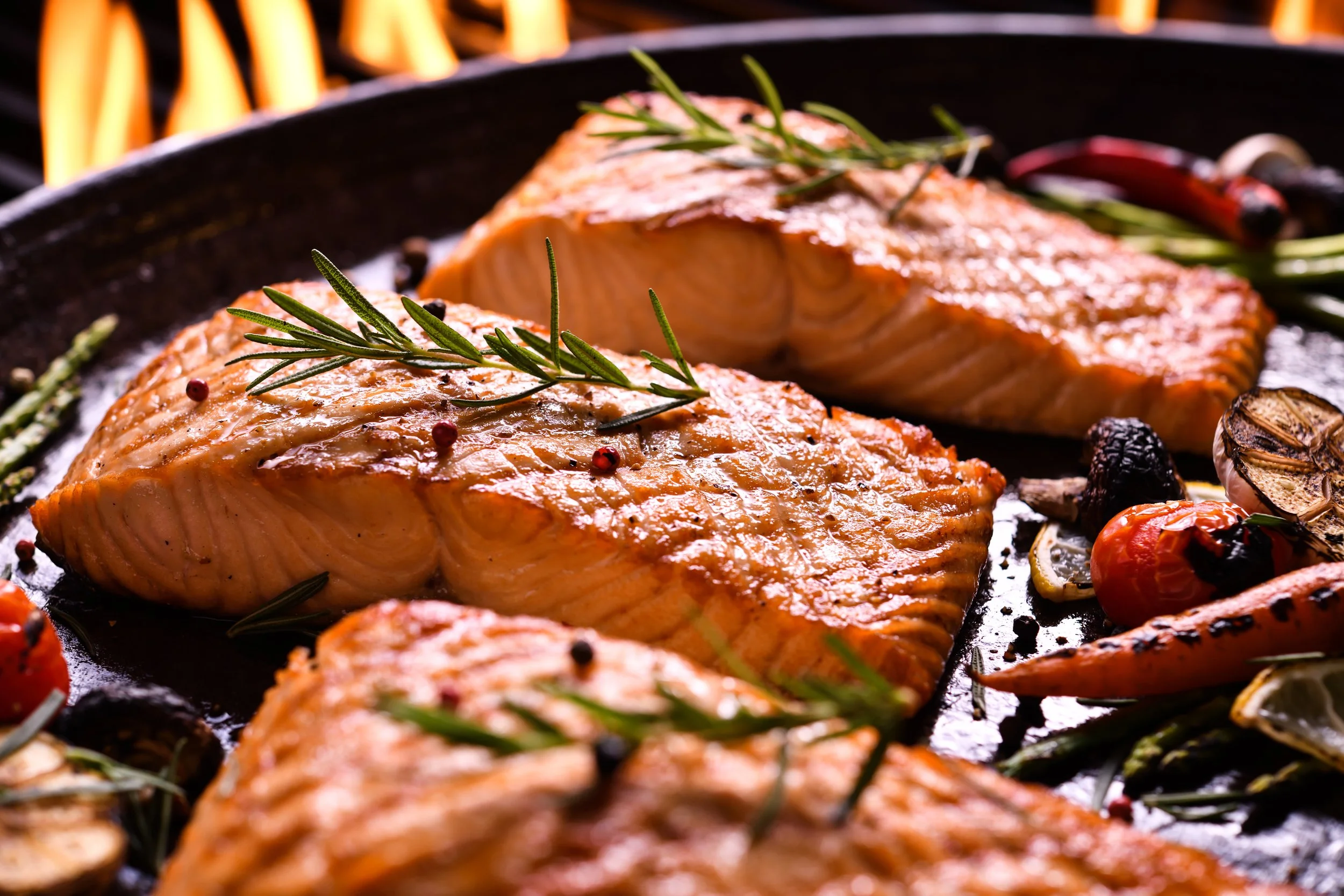 Salmon fillets on a grill with rosemary sprigs, peppercorns, cherry tomatoes, garlic, and chili peppers.