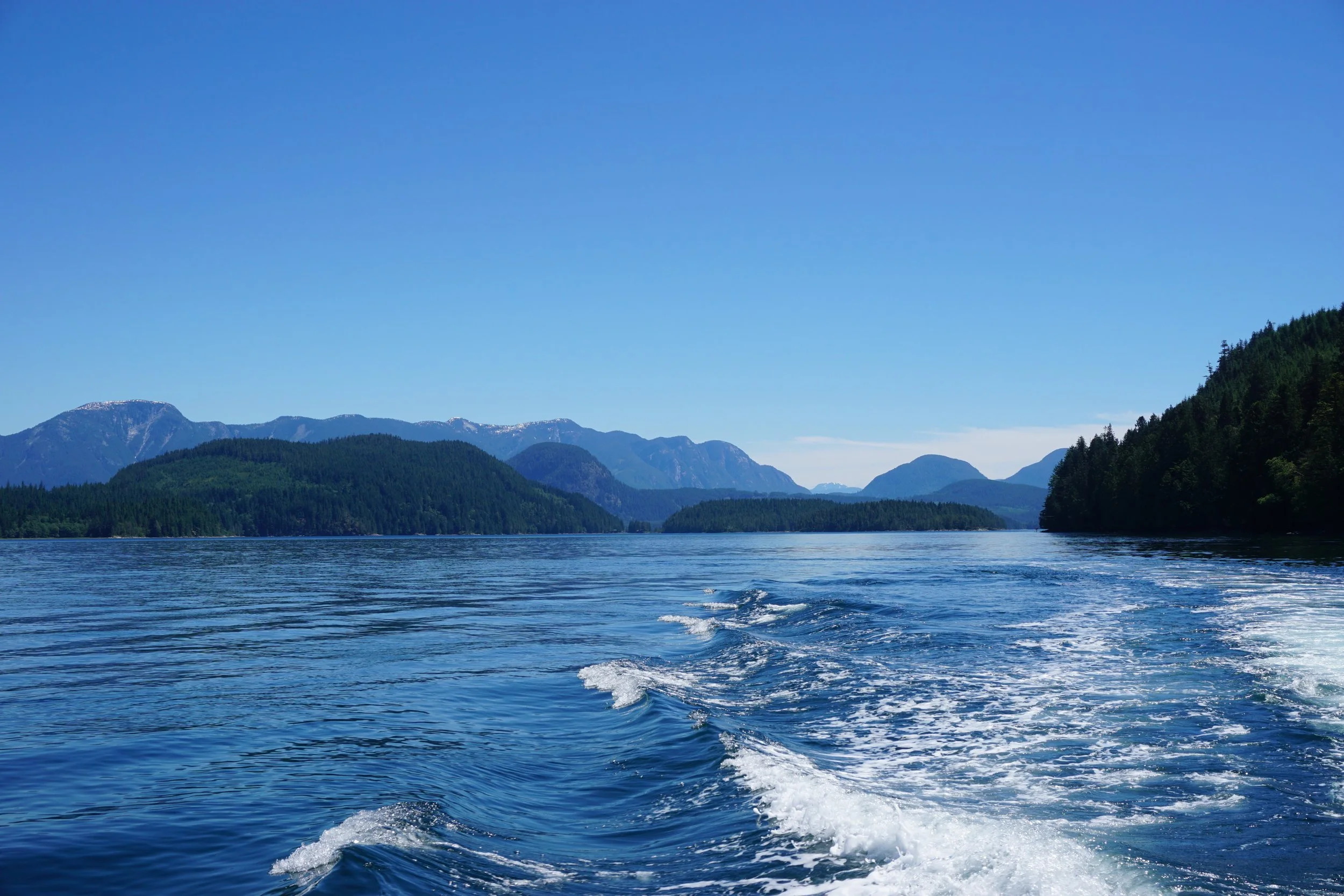 A body of water with visible wake from a boat, surrounded by forested mountains under a clear blue sky.