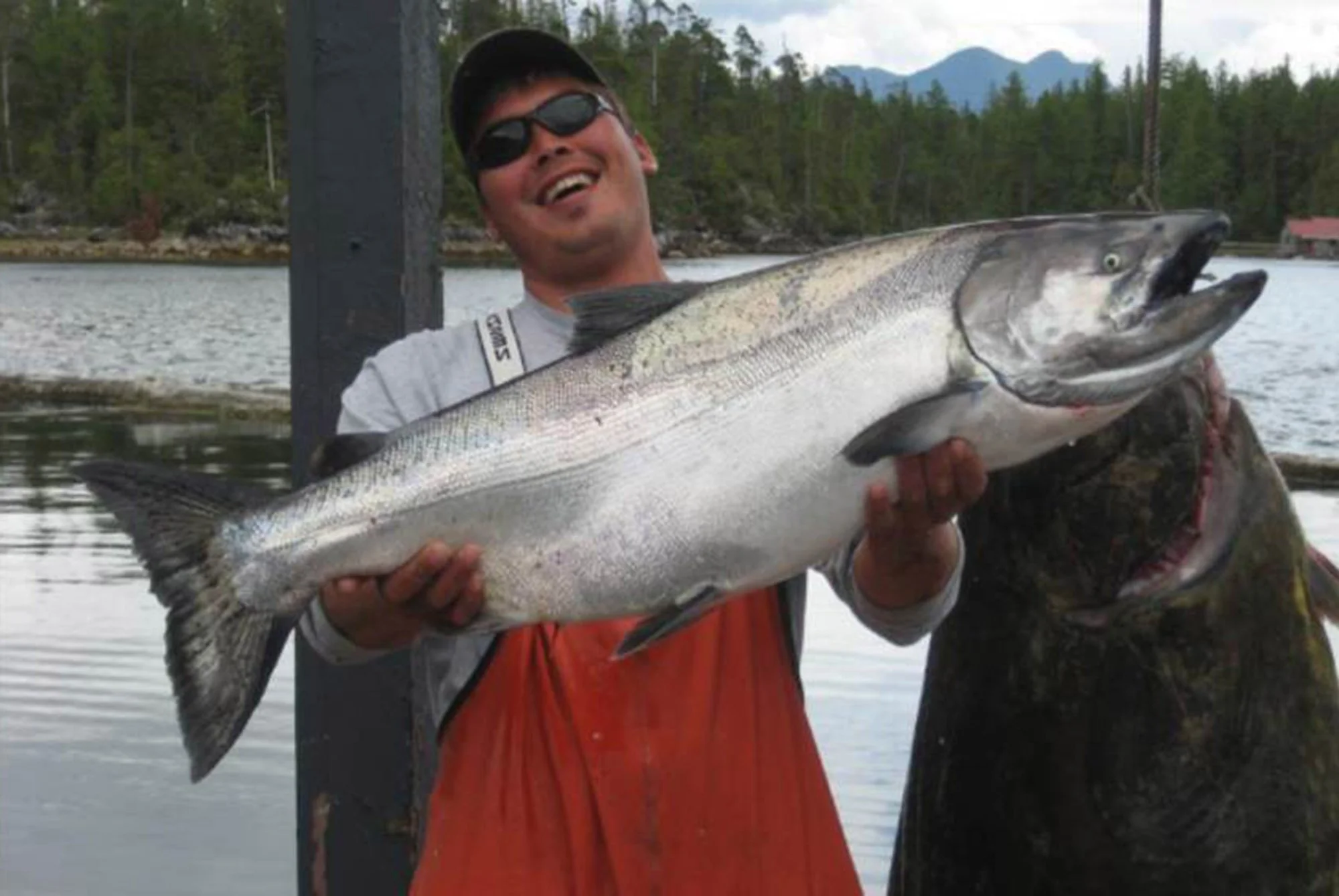 Man smiling and holding a large fish on a boat with water, trees, and mountains in the background.
