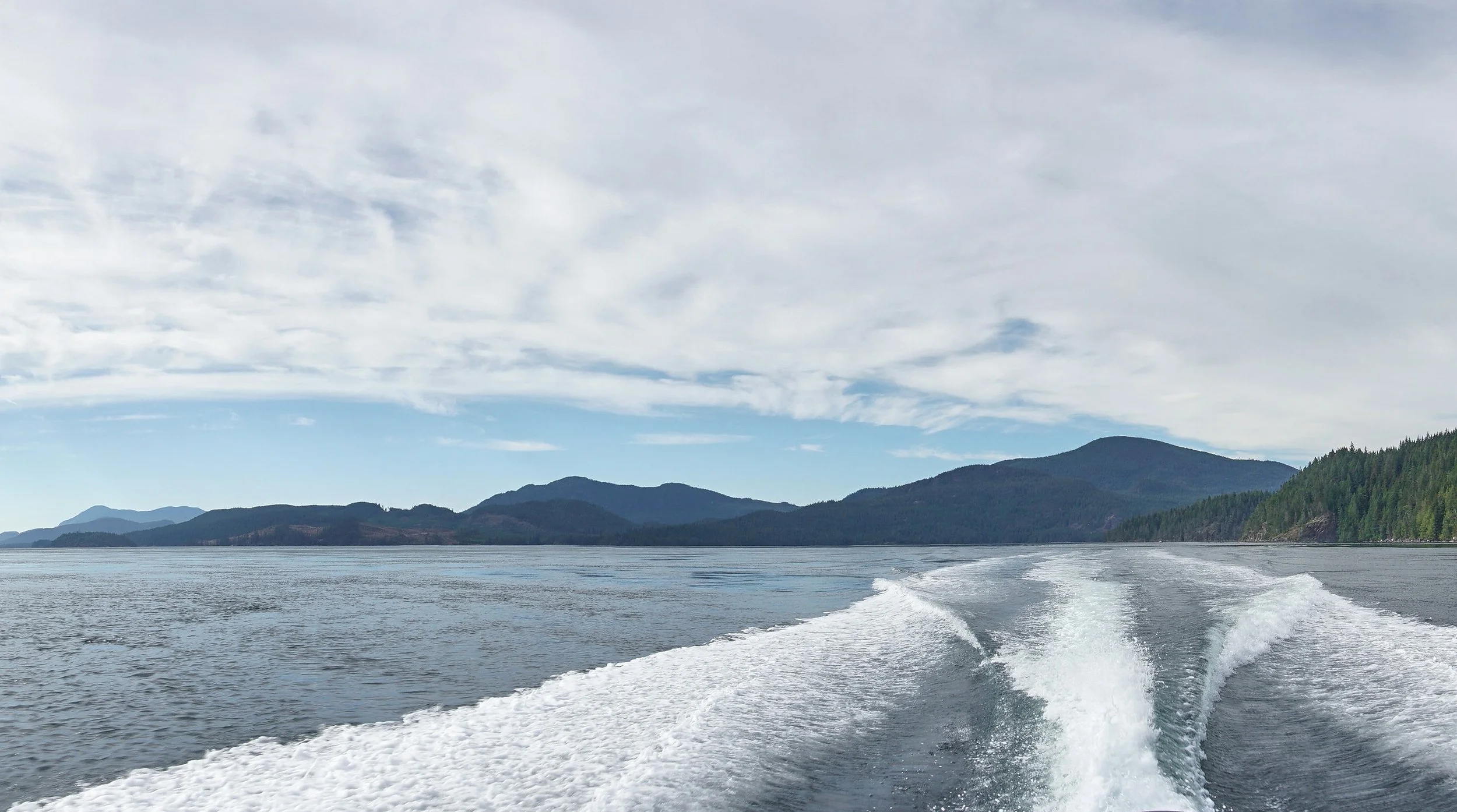 A view from a boat showing a wake in the water with distant mountains and a partly cloudy sky.