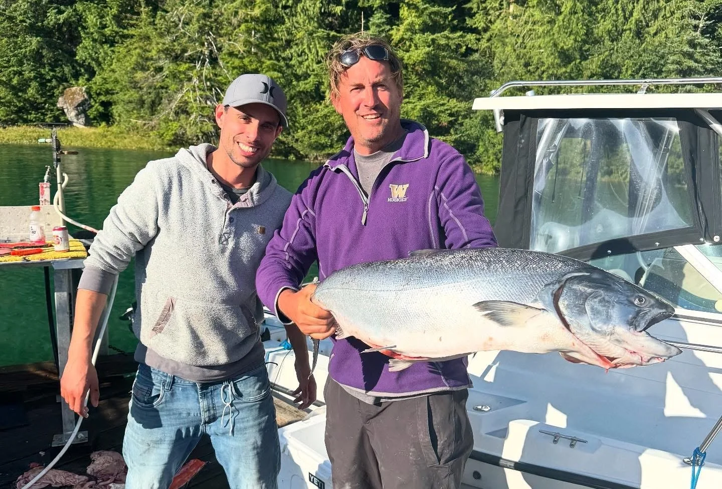 Two men on a boat holding a large fish, with a lake and trees in the background.