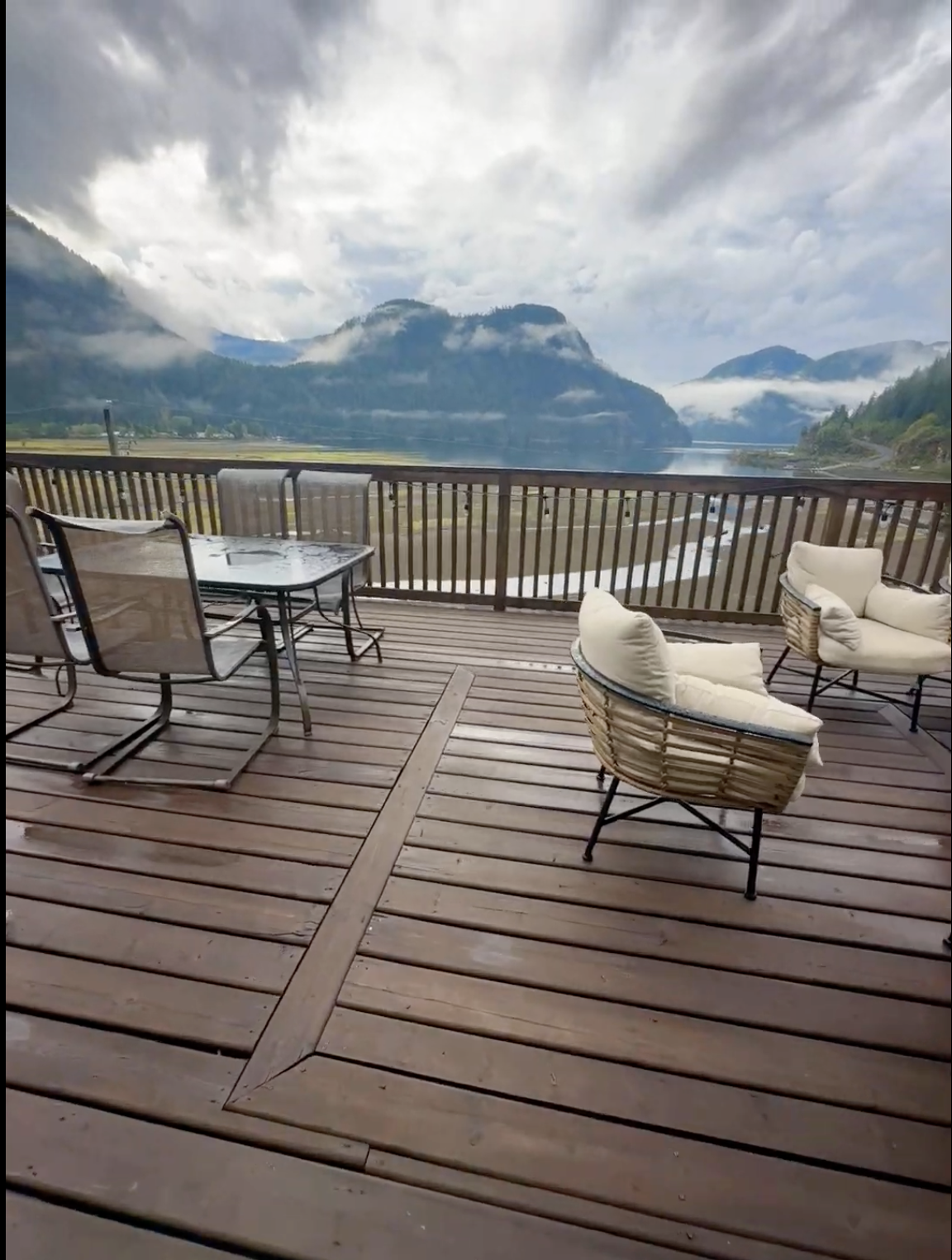 Balcony with outdoor chairs and table overlooking a river, mountains, and cloudy sky.