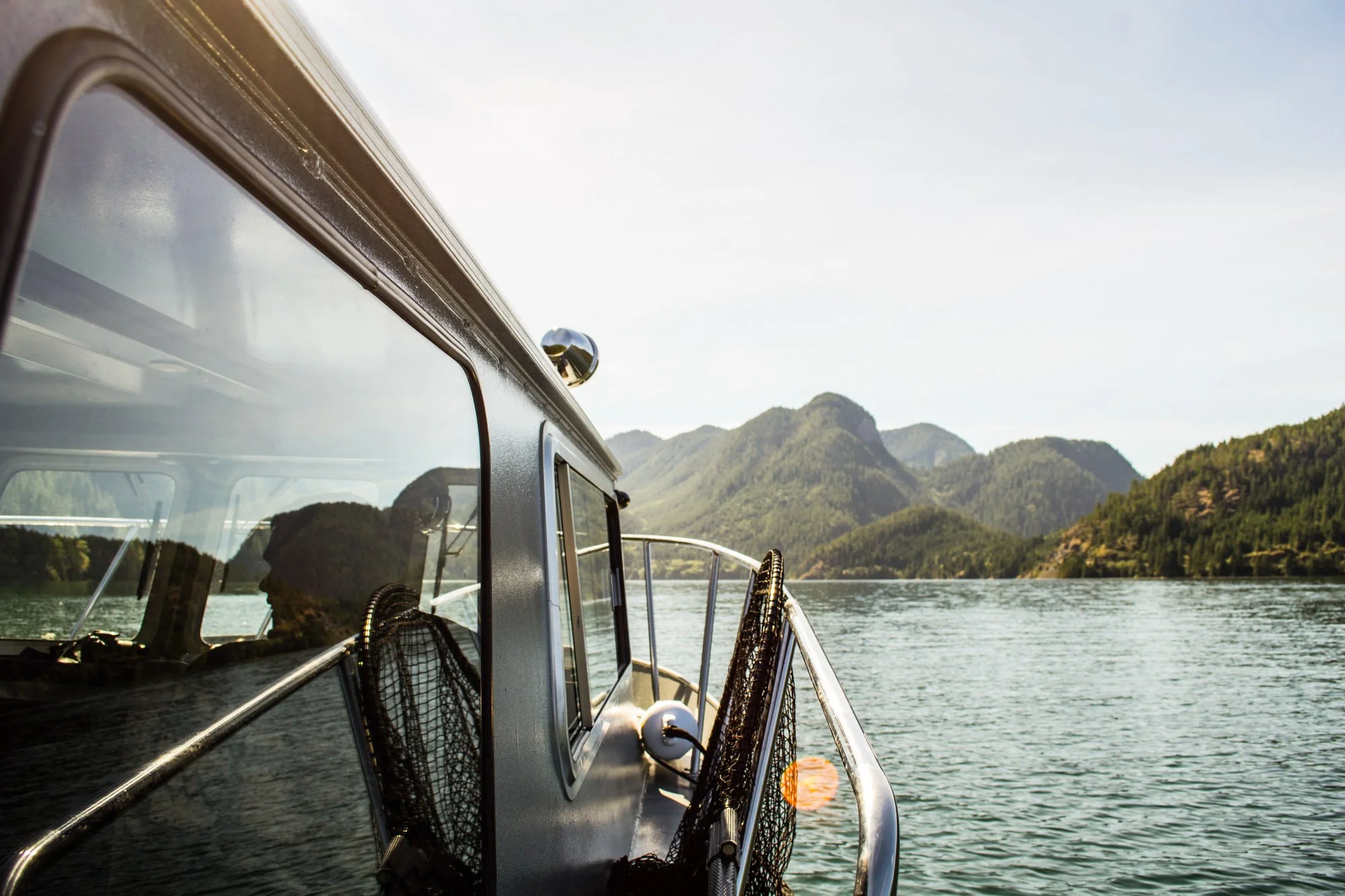 View from a boat on the water, showing boat's side with windows and netted storage compartments, with mountains and partly cloudy sky in the background.