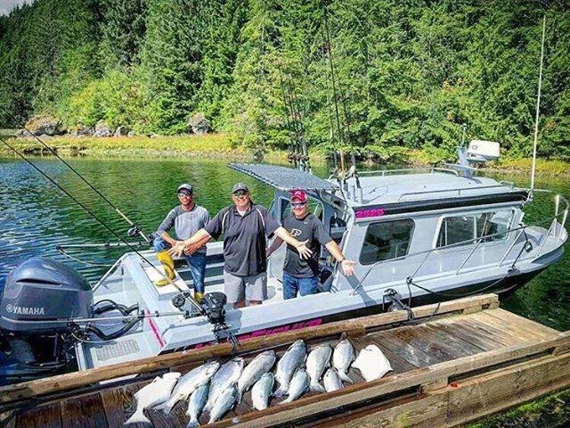 Three men standing on a boat docked at a wooden pier with fish laid out in front of them, surrounded by calm water and a densely forested shoreline in the background.