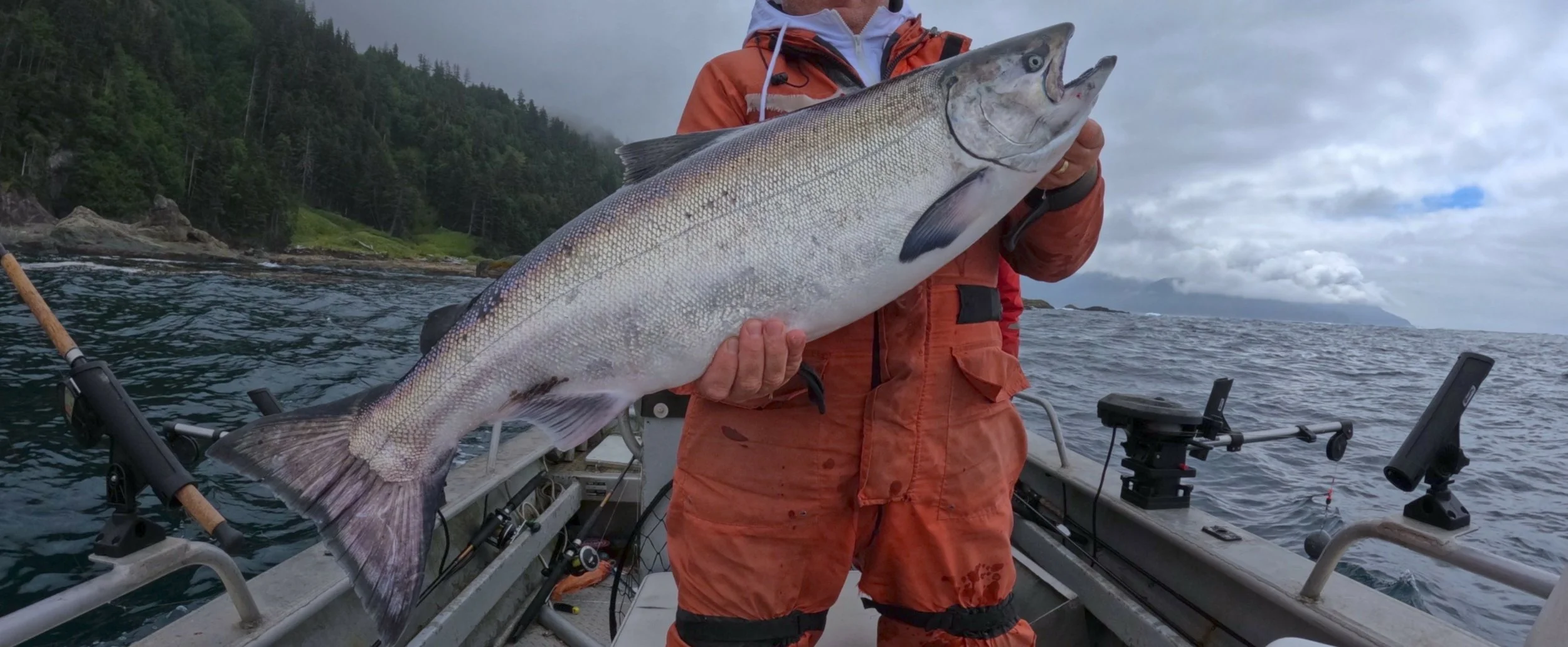 Person in an orange jacket holding a large fish on a boat in the water with a forested shoreline and cloudy sky in the background.