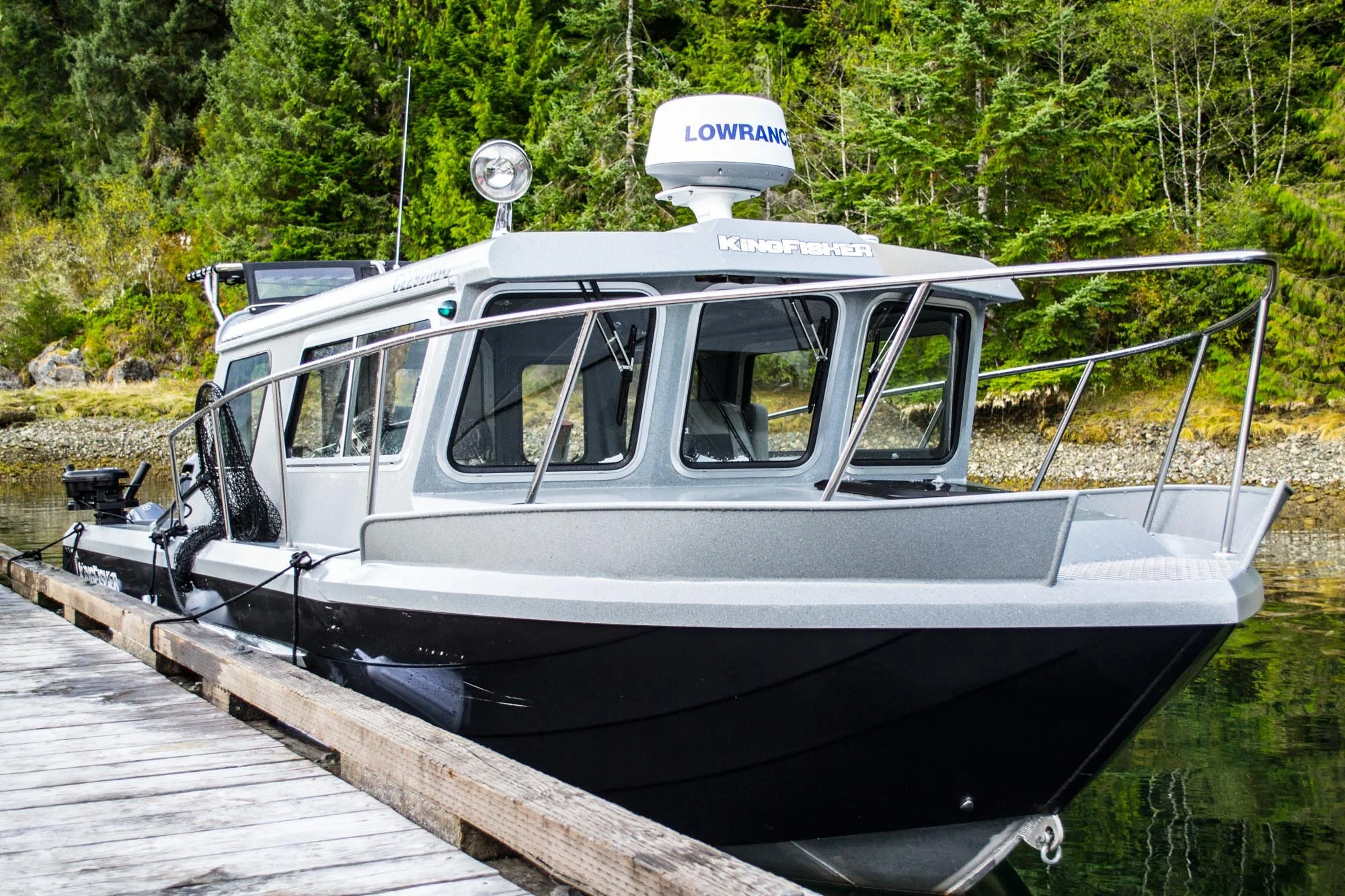 A white fishing boat with navigation equipment, including a Lowrance radar, is docked at a wooden pier in a green forested area.