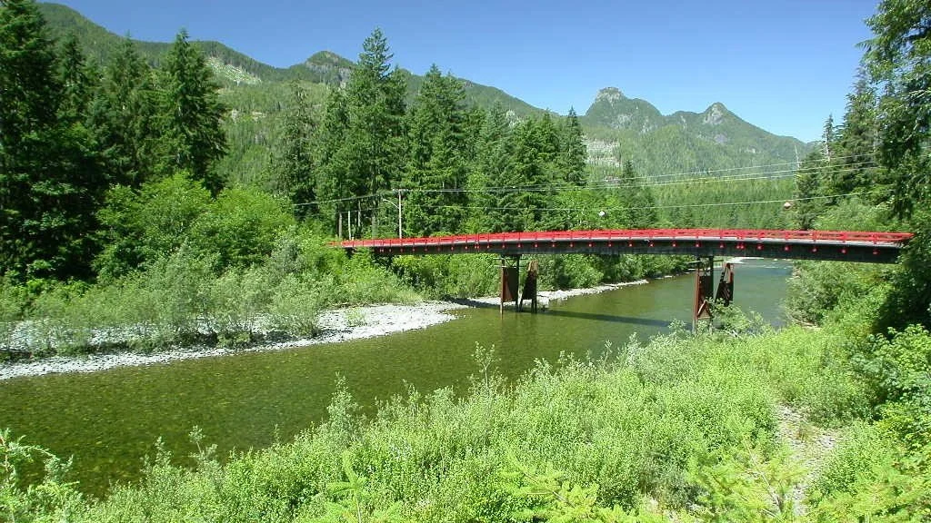 Red bridge crosses over a river surrounded by dense green trees and mountains in the background.