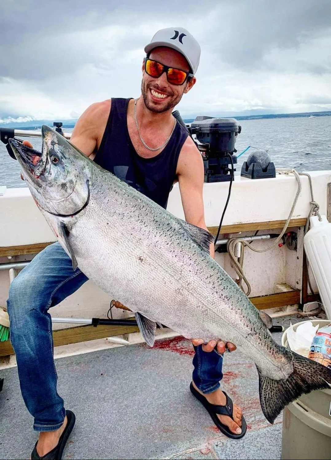 A man in sunglasses, a gray hat, and a sleeveless shirt is on a boat holding a large fish, with water and cloudy sky in the background.