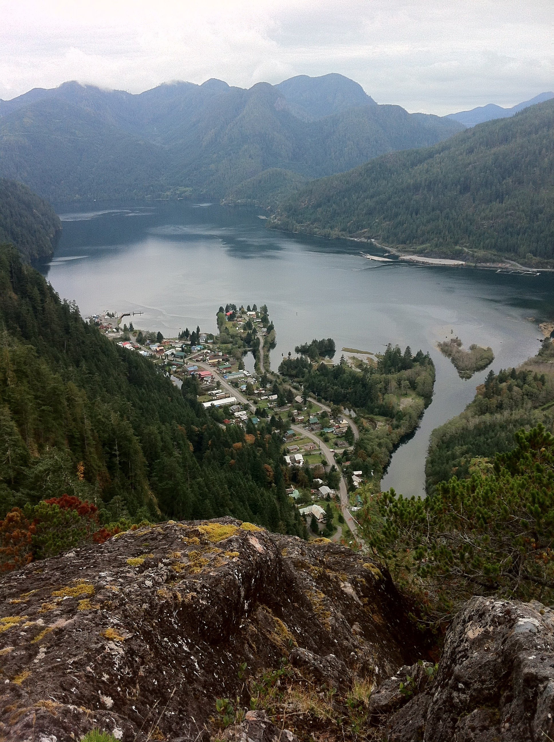 A view of a river with surrounding forested mountains, a small town at the river's edge, and overcast skies.
