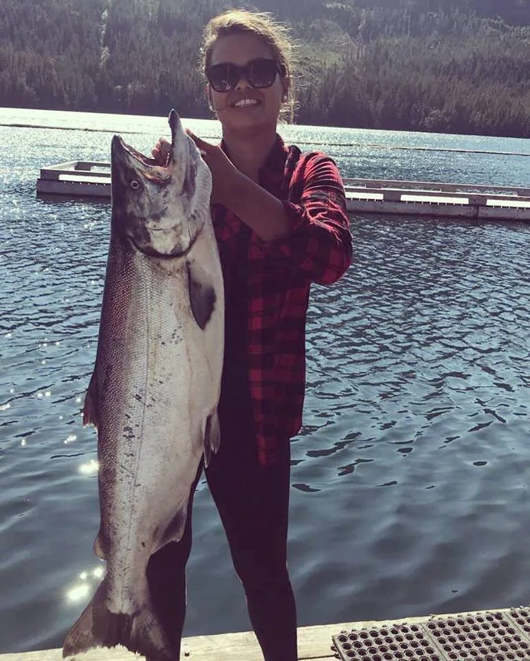 Woman with sunglasses in a red and black checkered shirt holding a large fish she caught, standing on a dock near a lake with distant mountains in the background.
