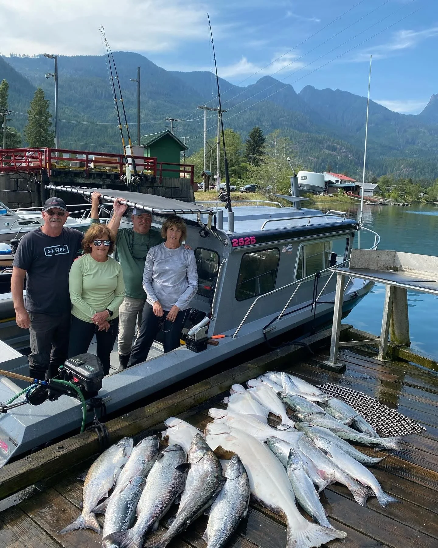 Four people standing on a dock by a boat with caught fish laid out on the dock, mountains in the background, and a body of water.