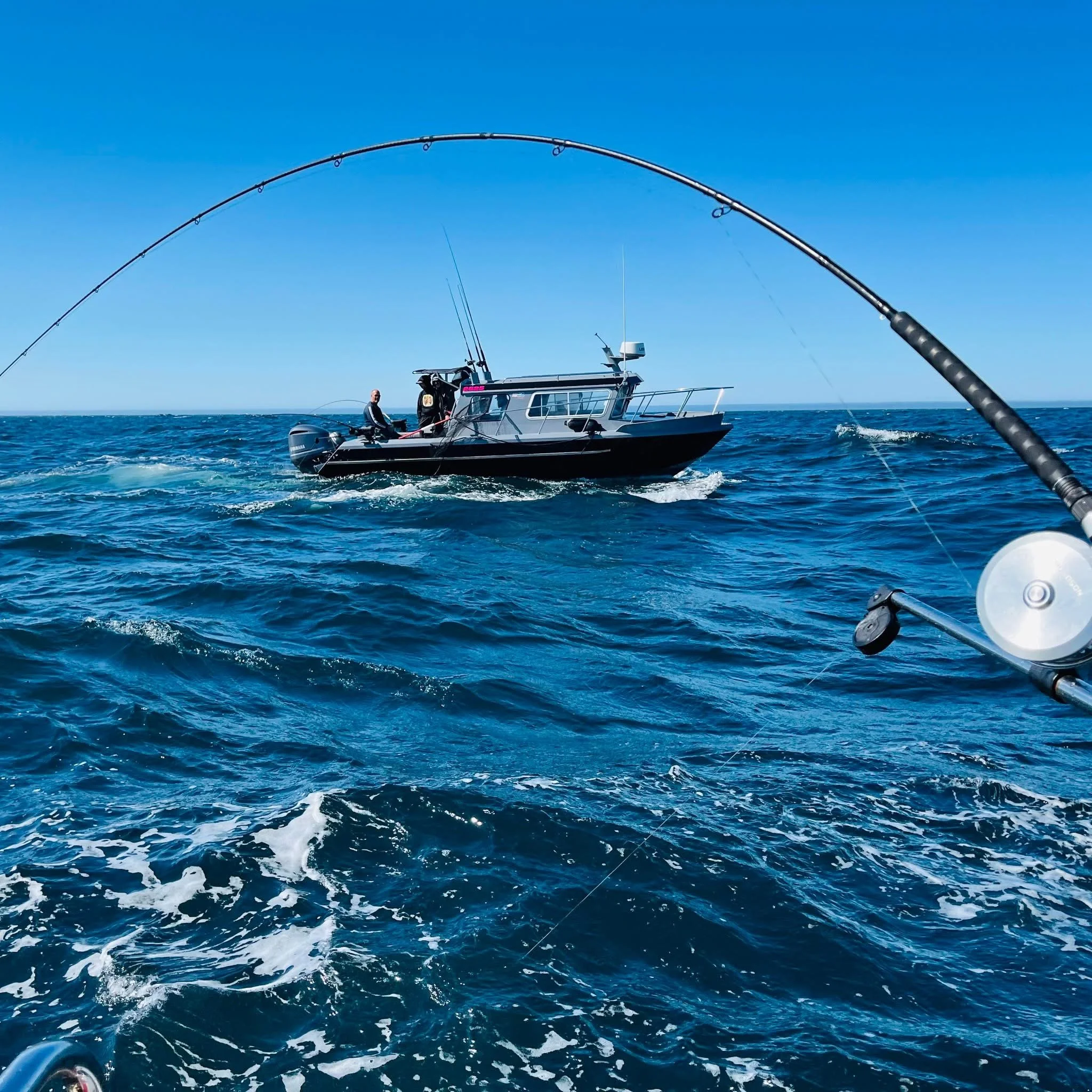 A fishing boat on the ocean with a person sitting on the stern, fishing rods extending over the water, and a clear blue sky above.