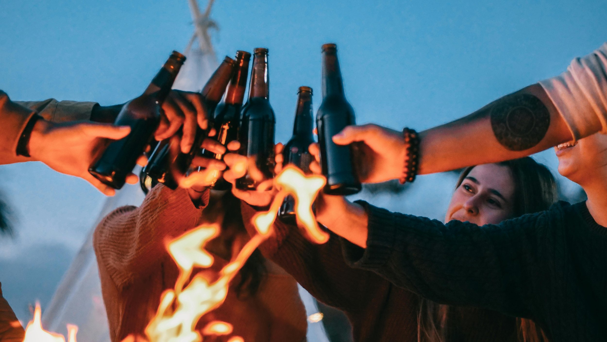 A group of people clinking beer bottles together outdoors with a fire in the foreground during evening.