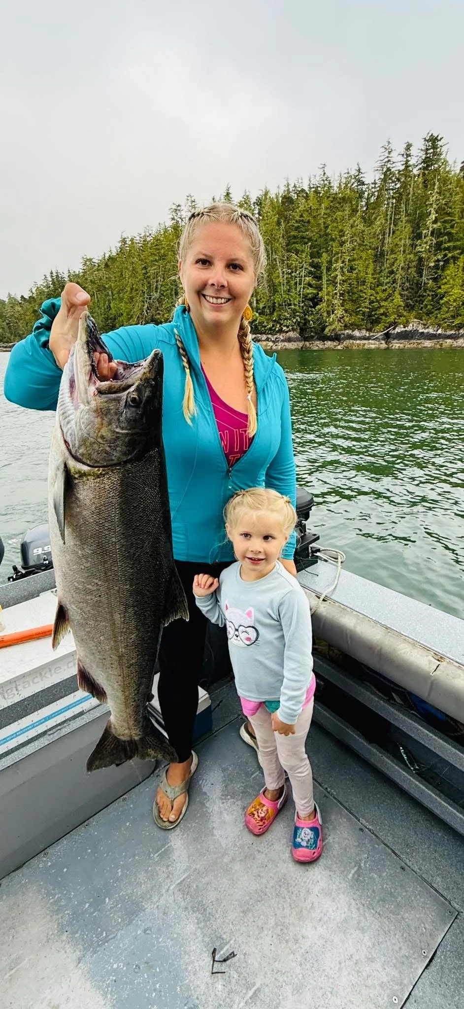A woman and a young girl on a boat, with the woman holding a large fish she caught. The woman has blonde hair in two braids and is wearing a blue jacket. The girl has blonde hair, wearing a gray shirt with a cat face, pink shorts, and pink shoes. The