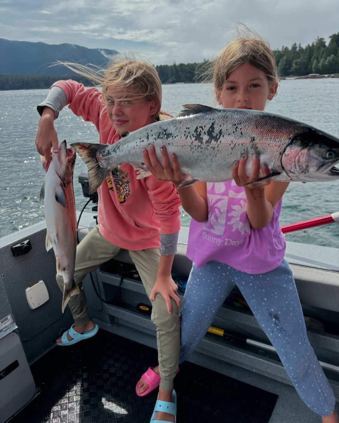 Two children on a boat holding large fish and a smaller fish, with a lake and mountains in the background.