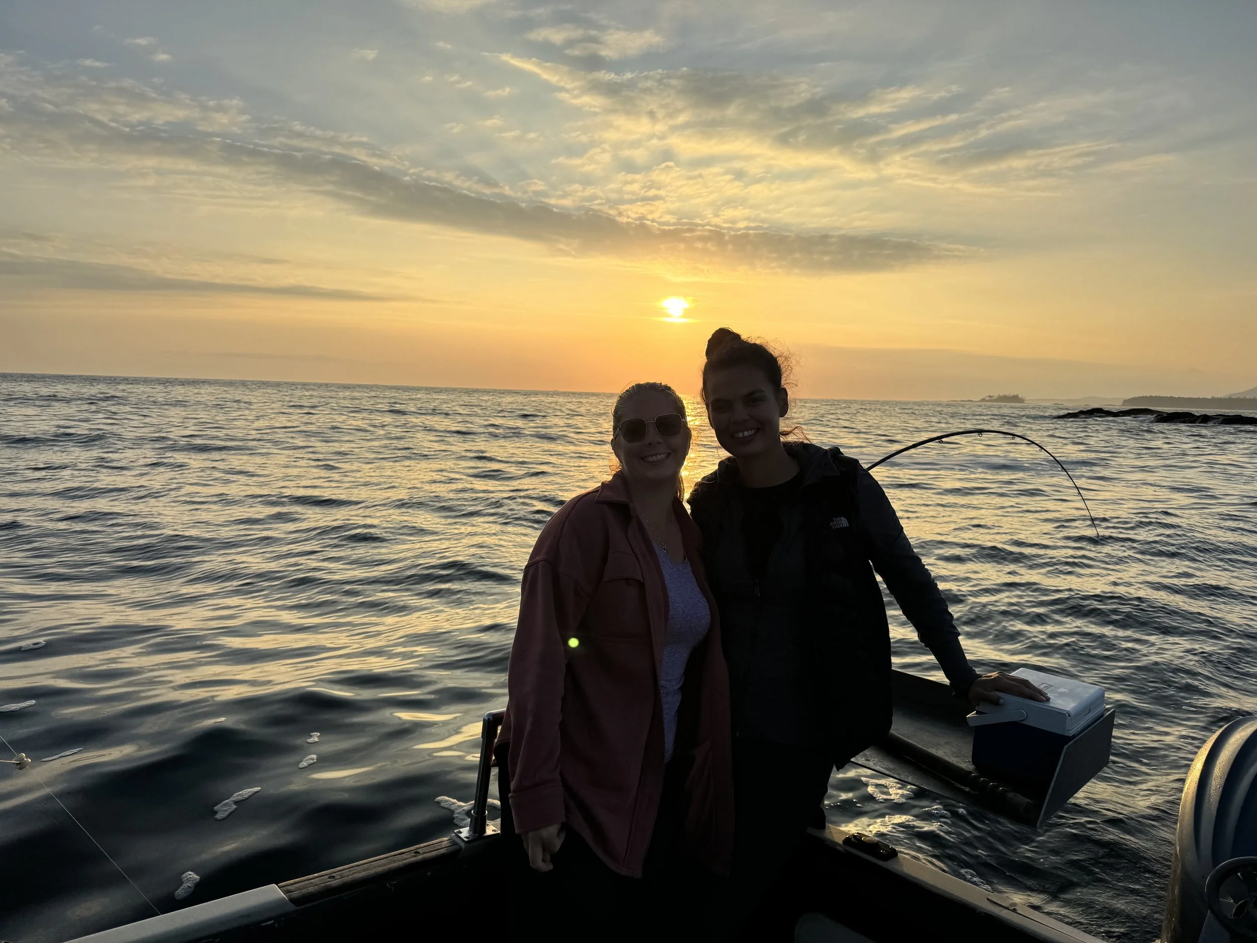 Two women smiling on a boat during sunset, with water and a partly cloudy sky in the background.