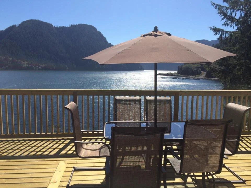 A wooden deck overlooking a lake with mountains in the background. There is a table with six chairs and a large patio umbrella providing shade.