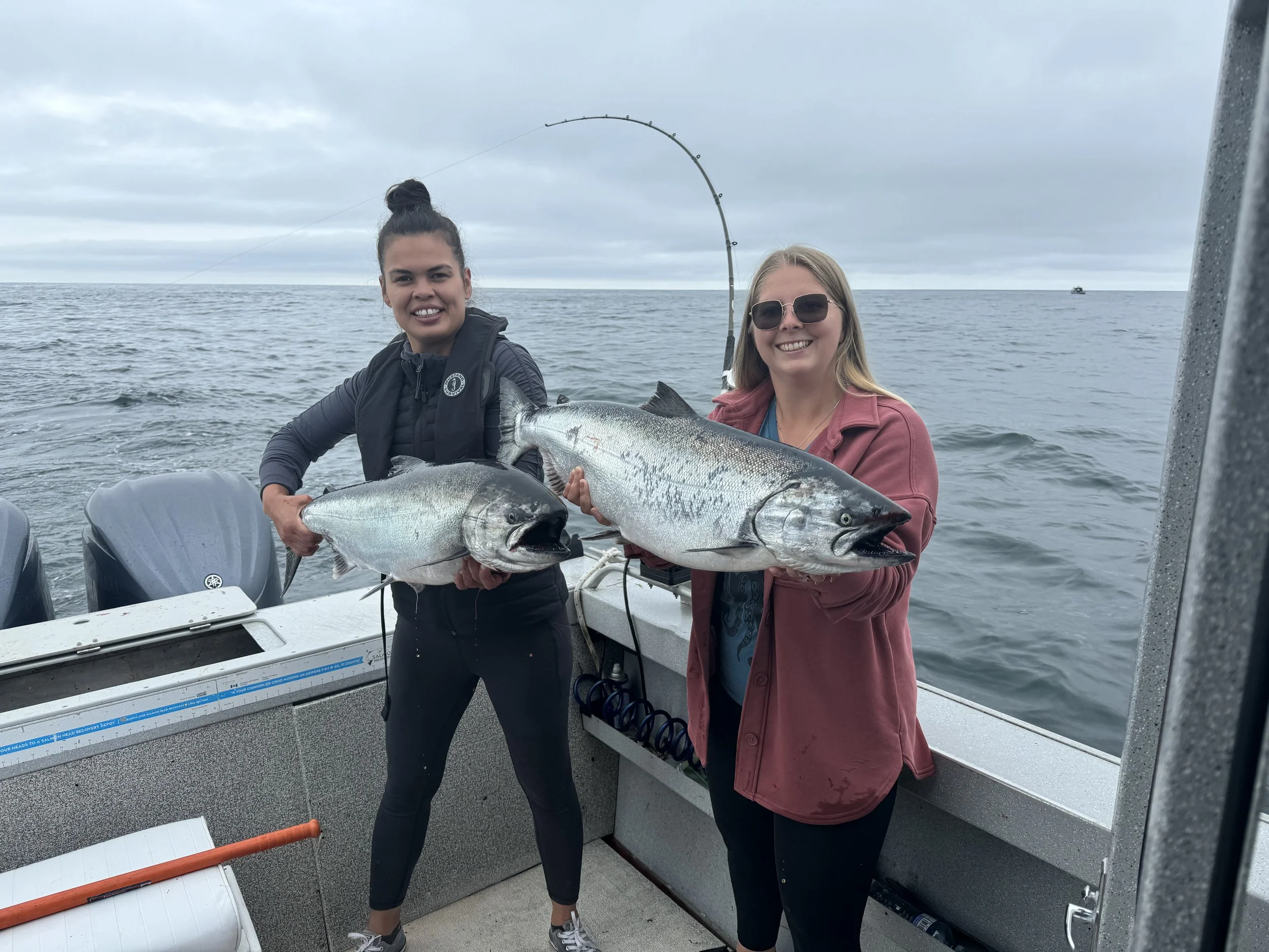 Two women on a boat holding large fish, smiling, with fishing rods in the background on an overcast day on the ocean.
