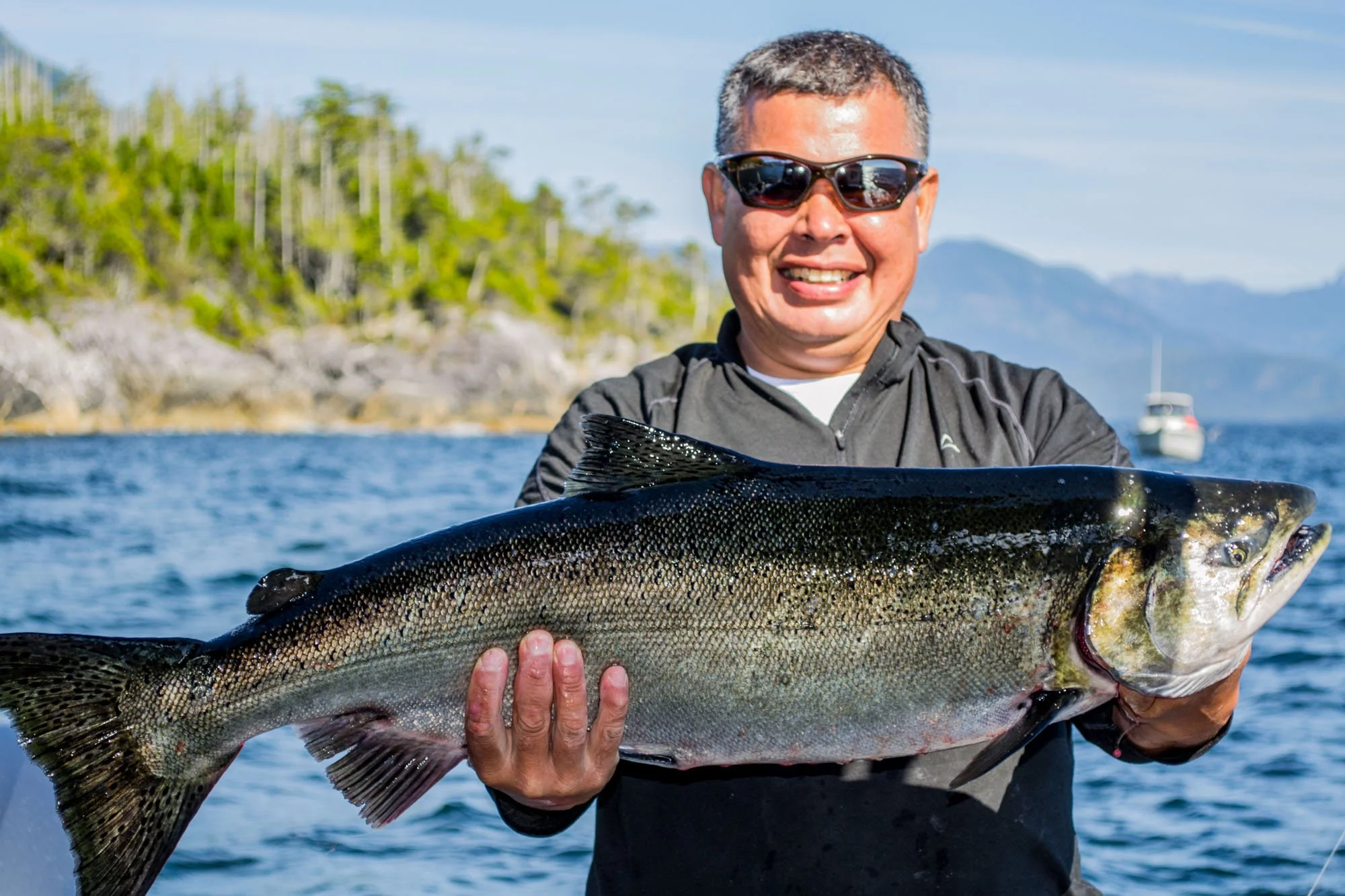 Man outdoors on a boat holding a large fish he caught, with trees, water, and a boat in the background.