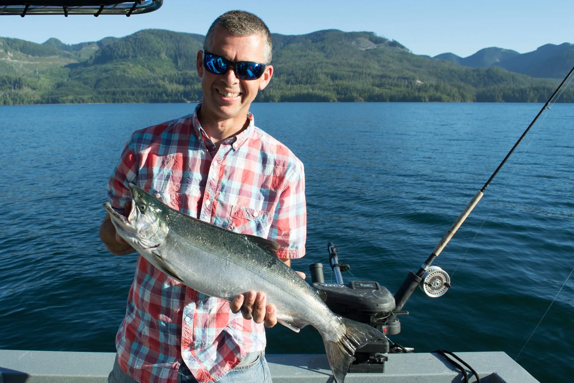 A man holding a large fish on a boat with fishing rods, water, and mountains in the background during daytime.