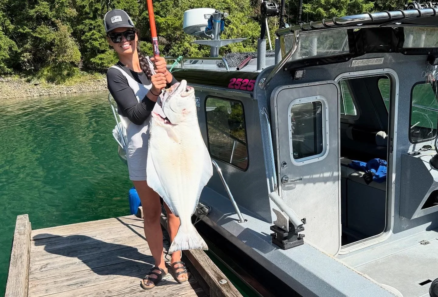 A woman on a dock holding a large fish next to a boat, smiling at the camera.