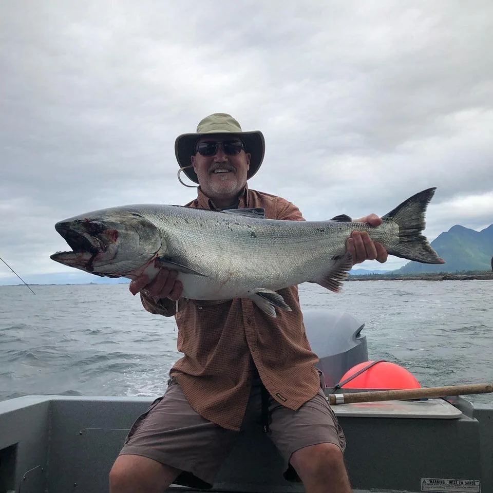 A man wearing sunglasses, a wide-brimmed hat, and outdoor clothing is smiling while holding a large fish on a boat in a body of water with mountainous scenery in the background.