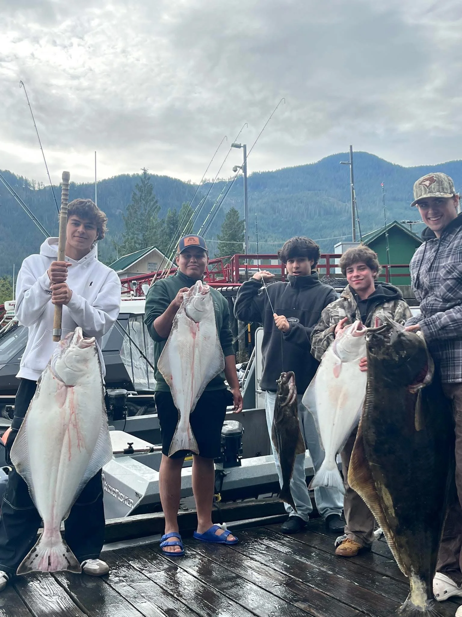 Group of five young boys standing on a boat dock holding large fish they caught during a fishing trip, with mountains, boats, and cloudy sky in the background.