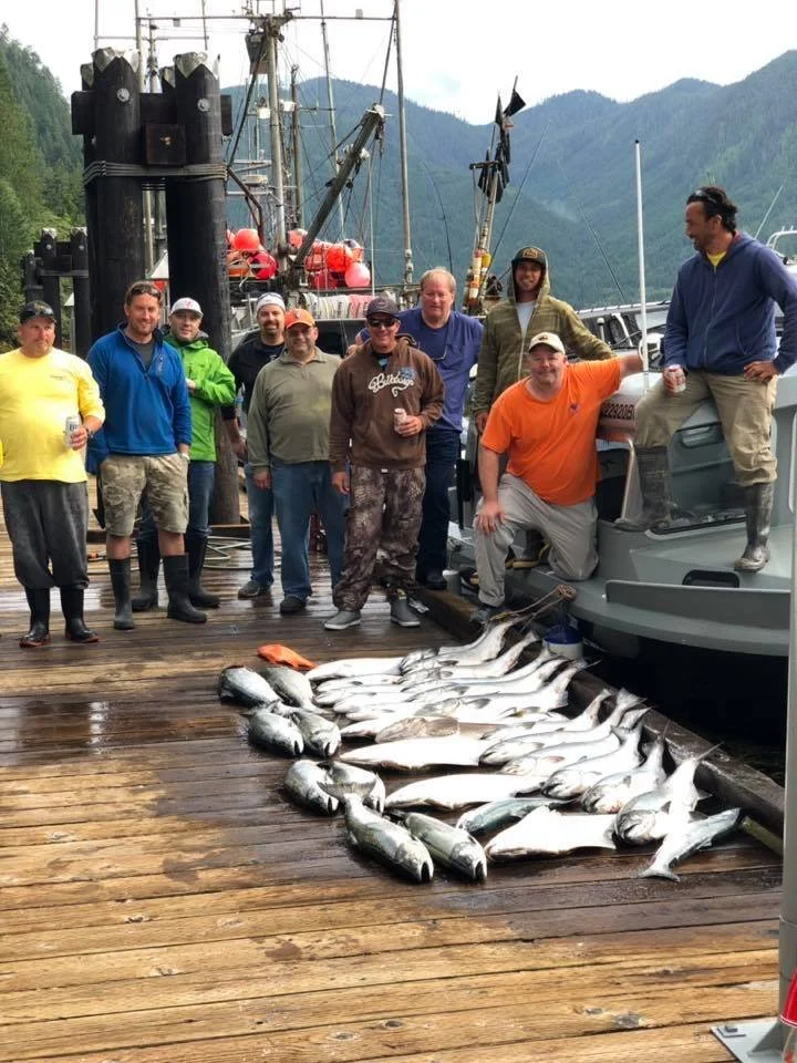 Group of men on a dock with a catch of fish, some large ships in the background, and a mountain range behind them.